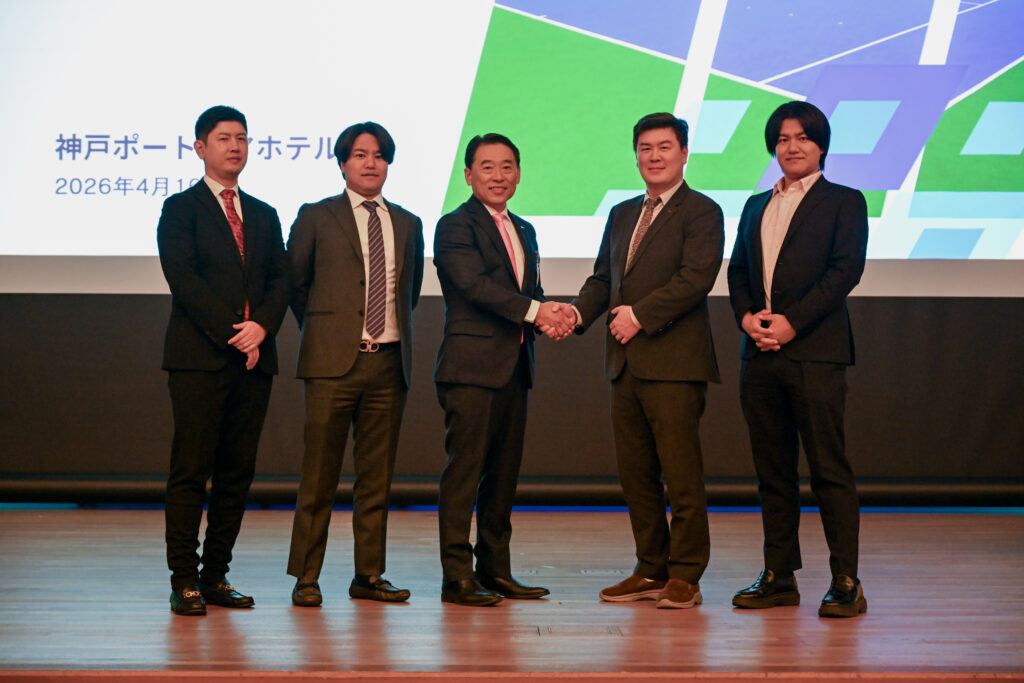 Five men in suits stand on a stage; two center figures shake hands while the others pose, with a bright geometric backdrop behind them.